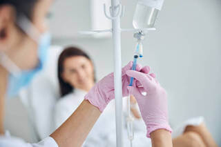 A nurse with pink gloves adjusts an IV drip while a patient rests in the background in a clinical setting.